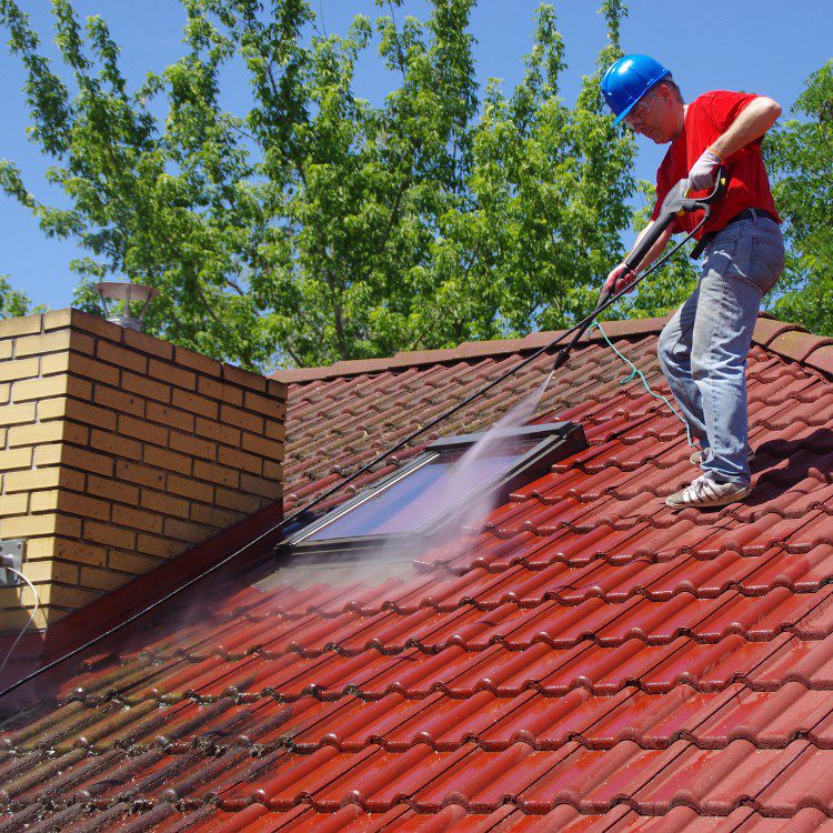 Worker in blue hard hat pressure washing red tile roof on sunny day